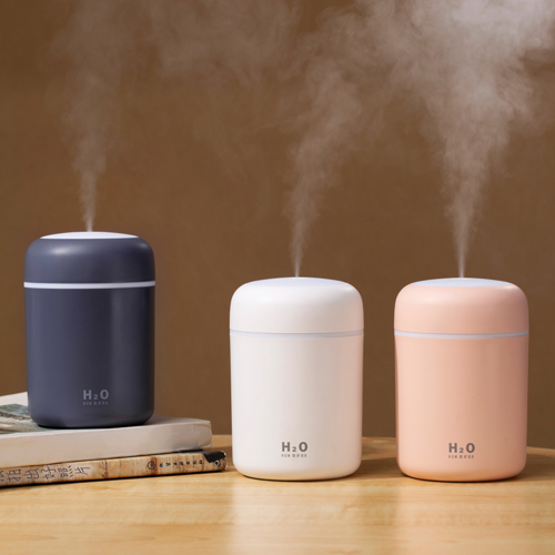 Three portable air humidifiers in navy blue, white, and peach colors, emitting mist, displayed on a wooden table.
