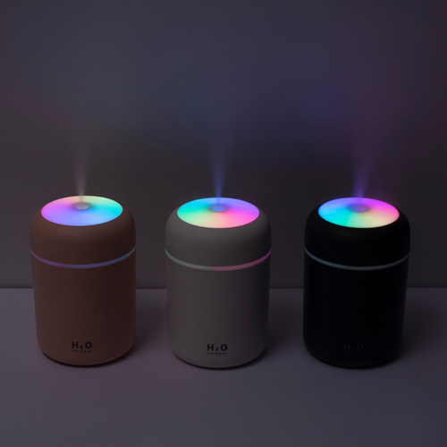 Three portable air humidifiers with colorful LED tops in pink, white, and black, emitting mist.