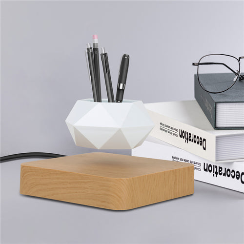 White geometric flowerpot levitating above a light wood base, holding pens, with books and glasses in the background.