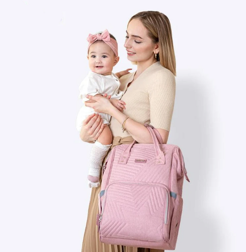 Modern Quilted Diaper Bag Backpack in soft pink being held by a smiling woman, while she carries a baby in her arms.